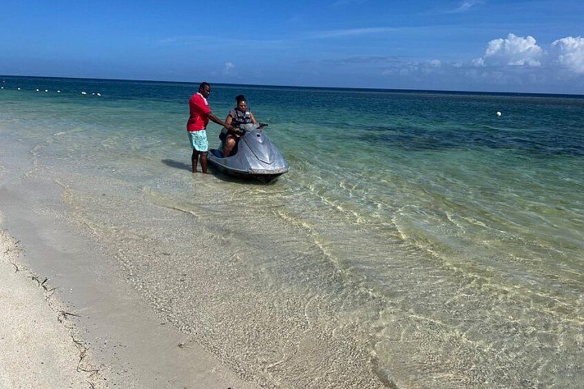 En jetski på en strand med klart, lavt vand, en person stående ved siden af den, og havet i baggrunden. 3