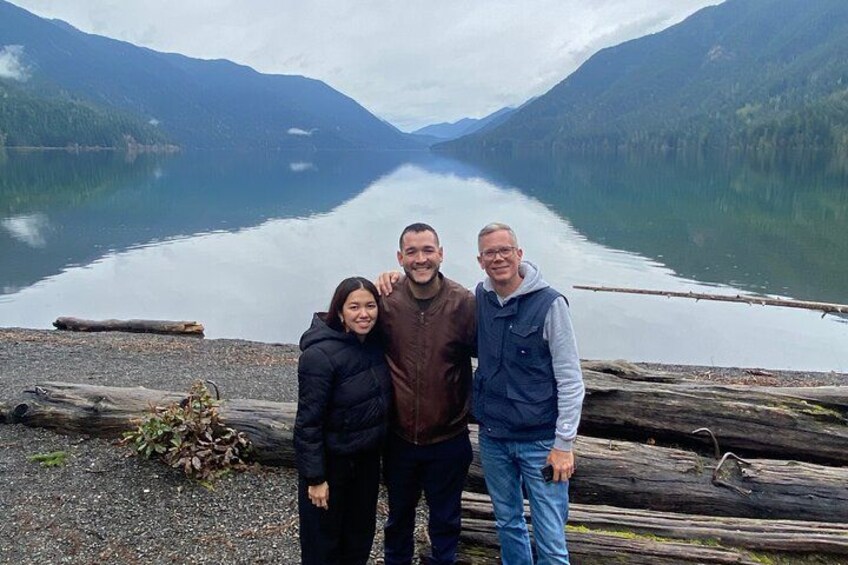 Three people standing by a calm lake with mountains in the background. 2