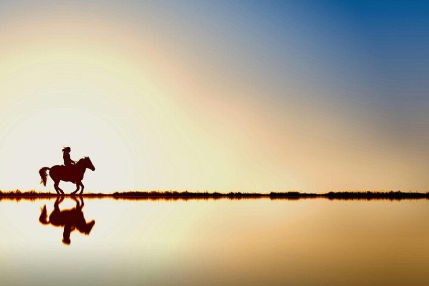 A silhouette of a person riding a horse on a calm body of water with a sunset in the background. 4
