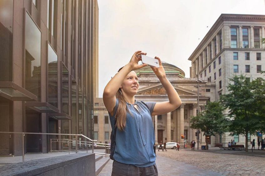 Eine Person fotografiert mit einem Smartphone in einer städtischen Umgebung mit modernen Gebäuden und Kopfsteinpflaster. 3