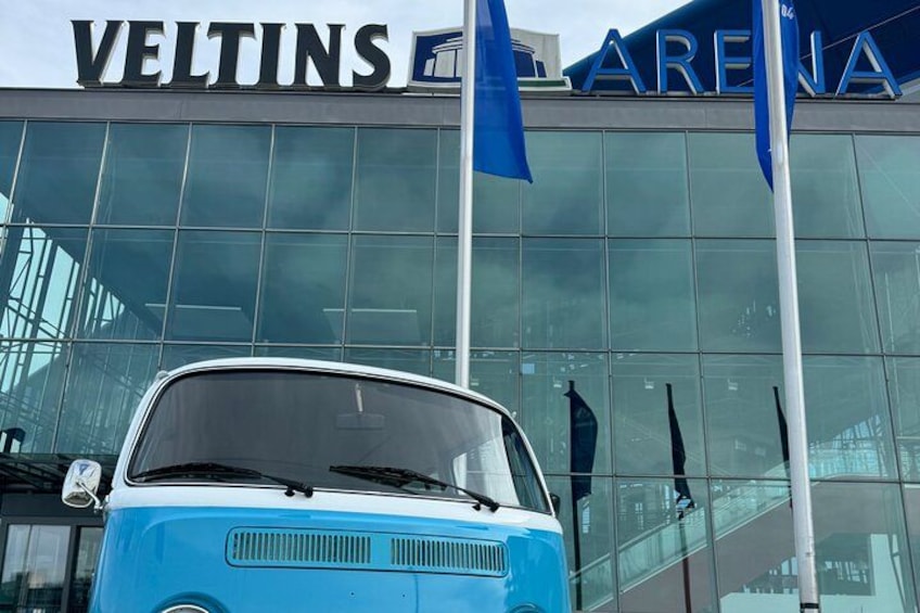 En blå bil foran en bygning med skilte for 'VELTINS ARENA'. 3