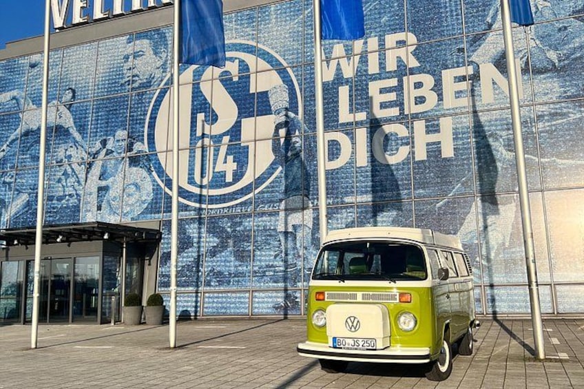 A vintage VW bus parked in front of a building with a large mural and flags. 1