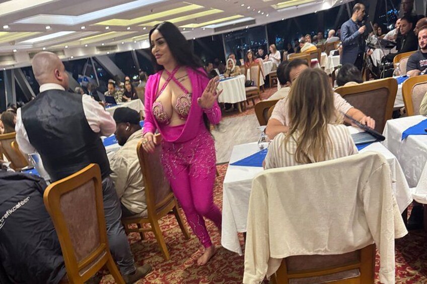 Une femme vêtue d’une tenue rose, debout dans un espace intérieur bondé où se trouvent des tables et des chaises. 2