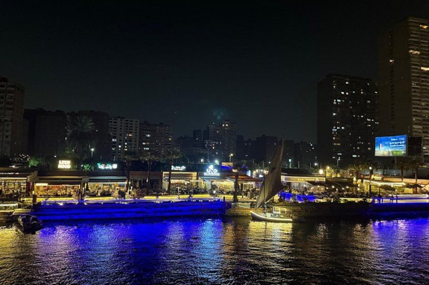 Een verlicht stadsbeeld bij nacht, met boten op een rivier, gebouwen op de achtergrond en een reclamebord. 4