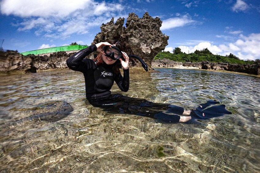 A person snorkeling in clear shallow water with rocky formations and a green structure in the background. 3