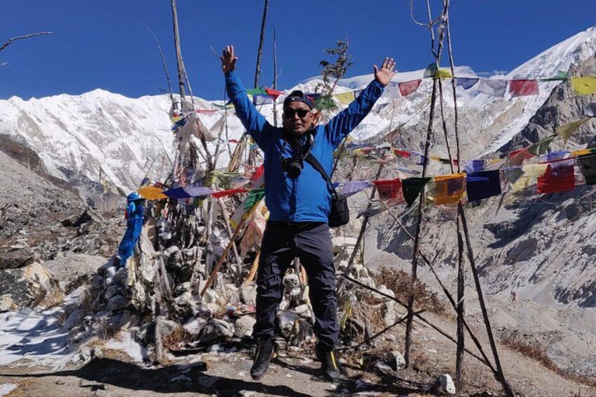 A person standing with arms raised in a mountainous area, surrounded by snow-covered peaks and colorful prayer flags. 1