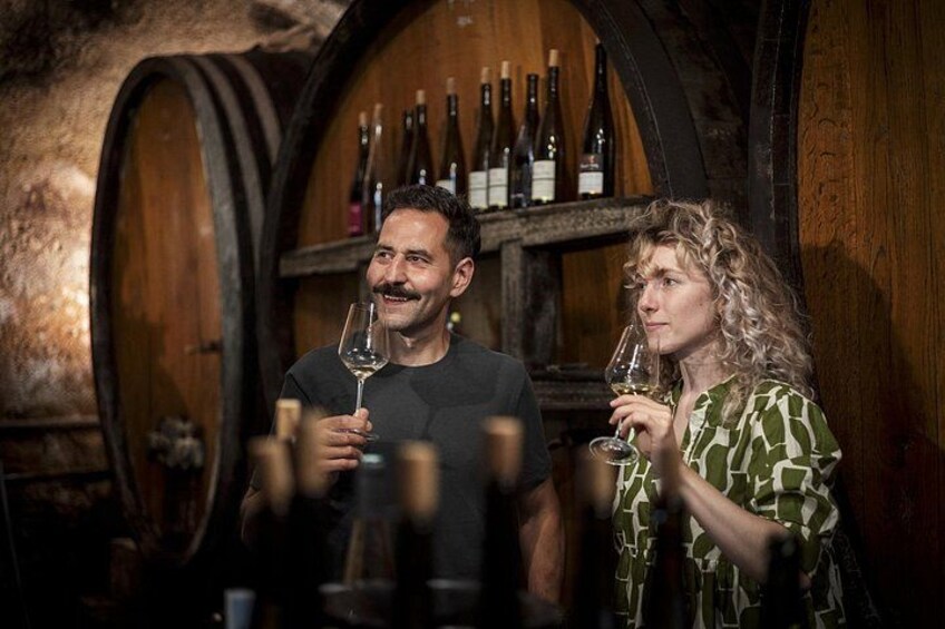 Two people holding wine glasses in a cellar with wooden barrels and wine bottles in the background. 4
