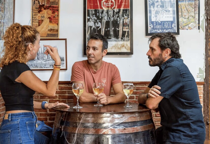 Three people sitting at a wooden table with glasses of wine, in a room with vintage posters on the wall. 4