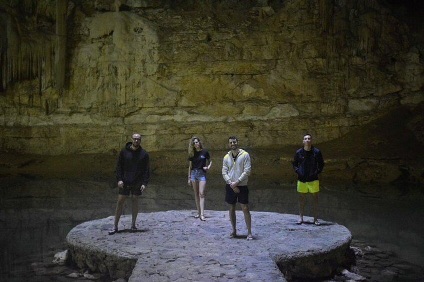 Four people standing on a circular stone platform in a cave with rock walls. 4