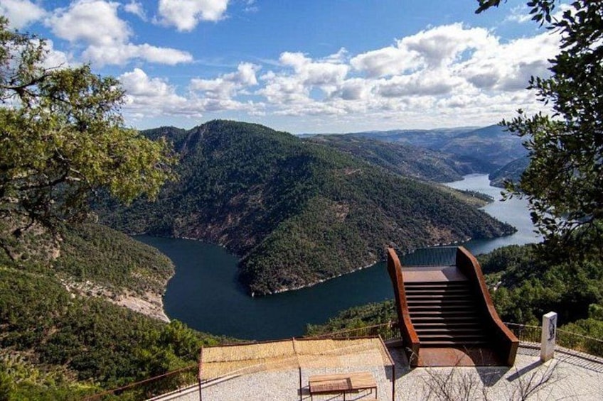 Un punto panoramico con una scala in legno che conduce a una panchina, con vista su un lago circondato da colline. 3