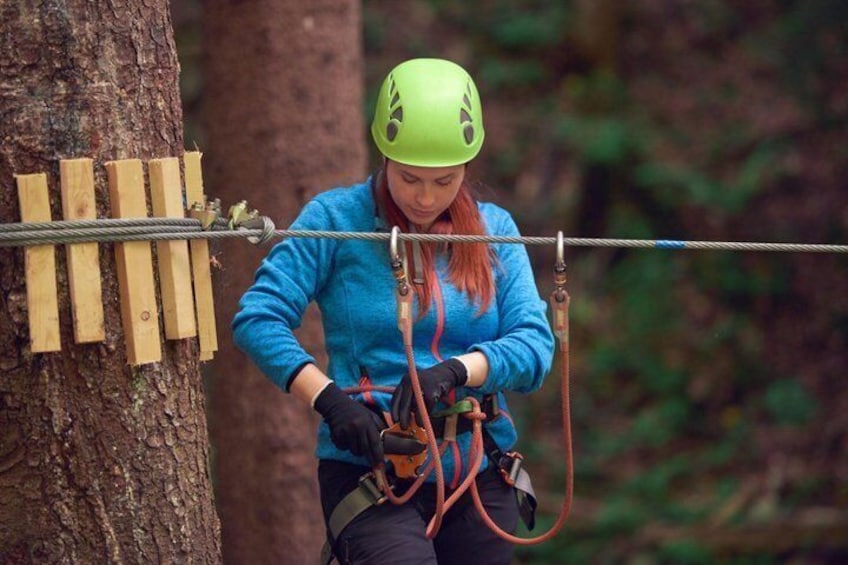 A person in a blue jacket and green helmet preparing for a zip-lining activity, with wooden planks and a cable attached to a tree. 1