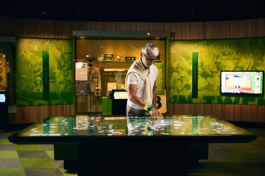 A man standing at a table with a green and yellow patterned wall behind him. 1