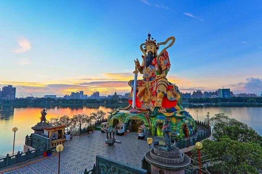 Una gran estatua de una divinidad sobre una plataforma, con un lago y el horizonte de la ciudad al fondo. 1