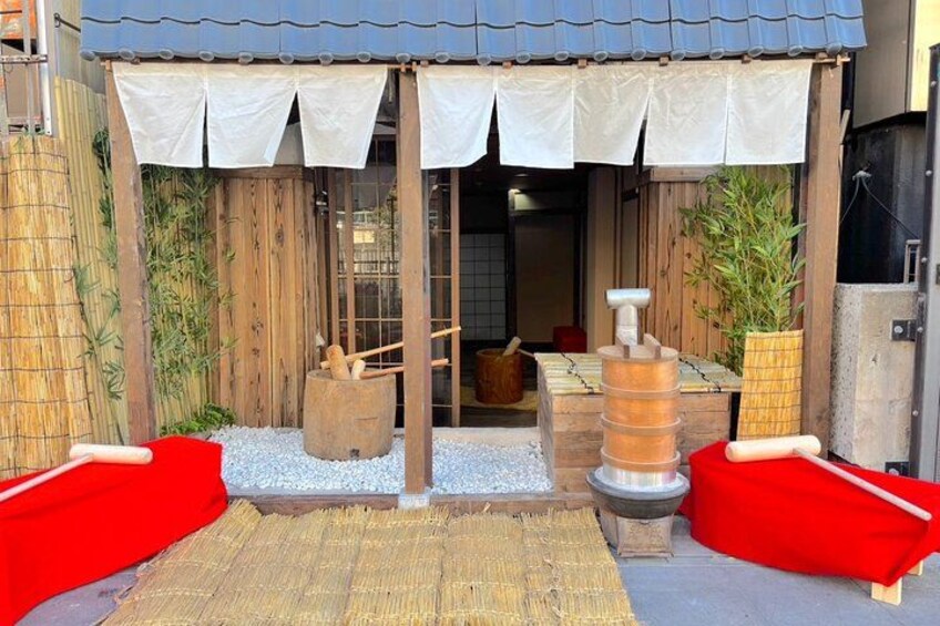 A traditional Japanese building with a wooden exterior, a tiled roof, and a covered entrance. There are red cushions outside, a stone basin, and a wooden barrel. 3