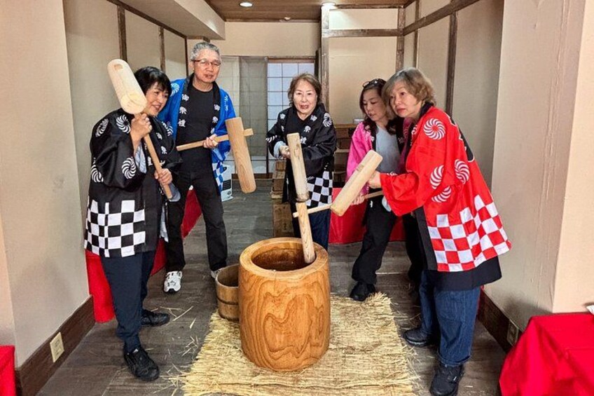 Un grupo de personas vestidas con atuendo tradicional sostienen mazos de madera, de pie en un pasillo con un barril grande de madera en el centro. 2