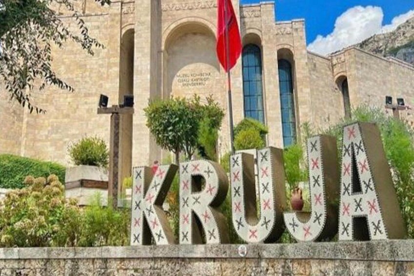 Ein Steinhaus mit dem Schriftzug 'KRUGA' in großen Lettern, einer roten Flagge und einer Steinmauer mit Grünpflanzen. 1