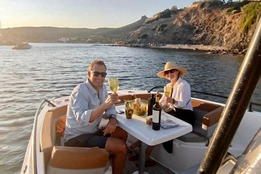 A boat with two people toasting with drinks, a table with bottles and glasses, and a rocky coastline in the background. 3