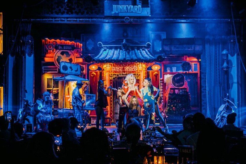 A neon-lit stage with performers, a sign reading 'JUNKYARD,' and a crowd of spectators. 1