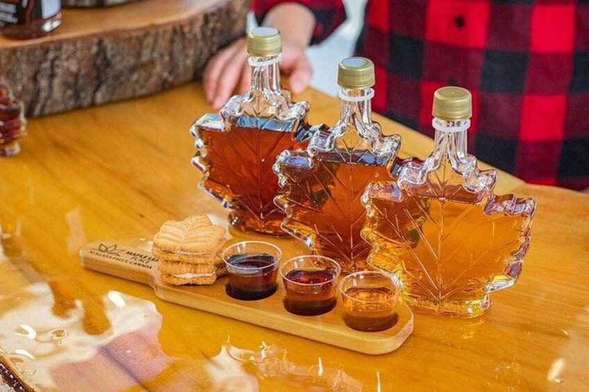 Une table en bois sur laquelle reposent trois carafes en verre remplies de sirop d’érable, un plateau en bois avec une bouteille de sirop d’érable et de petits récipients contenant du sirop. 3