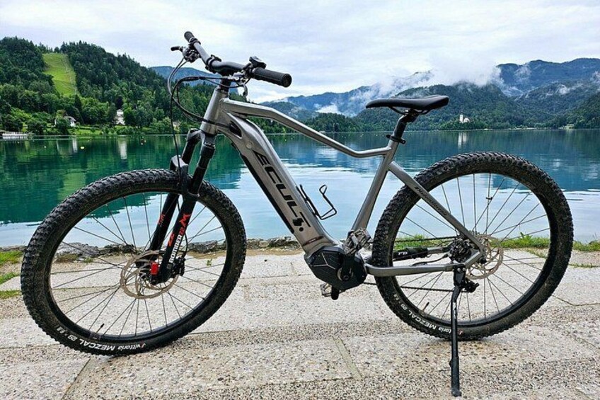 En mountainbike med en framgaffel med fjädring, en vattenflaska och ett bagageställ baktill, parkerad vid en sjö med berg i bakgrunden. 1