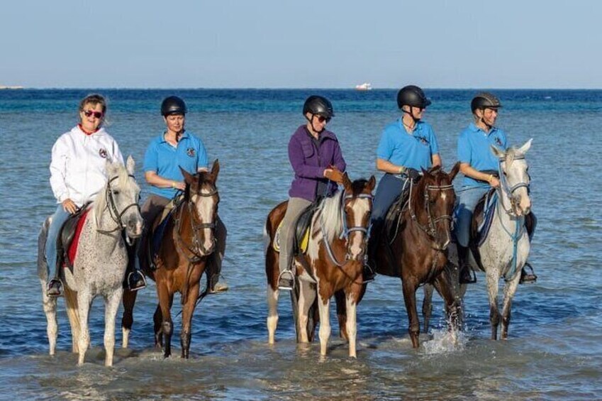Five people riding horses along a beach with the ocean in the background. 1