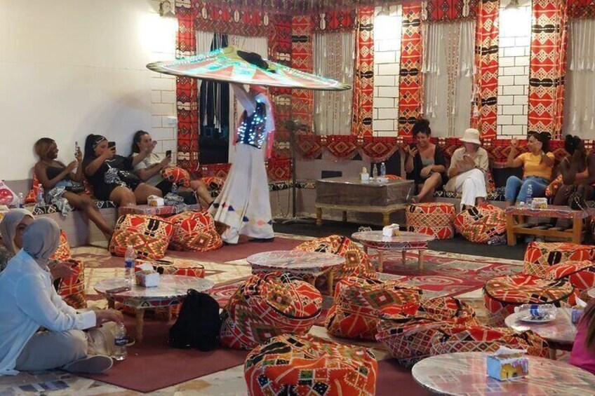 A group of people seated around tables in a decorated room with patterned cushions and a person holding a large umbrella. 3