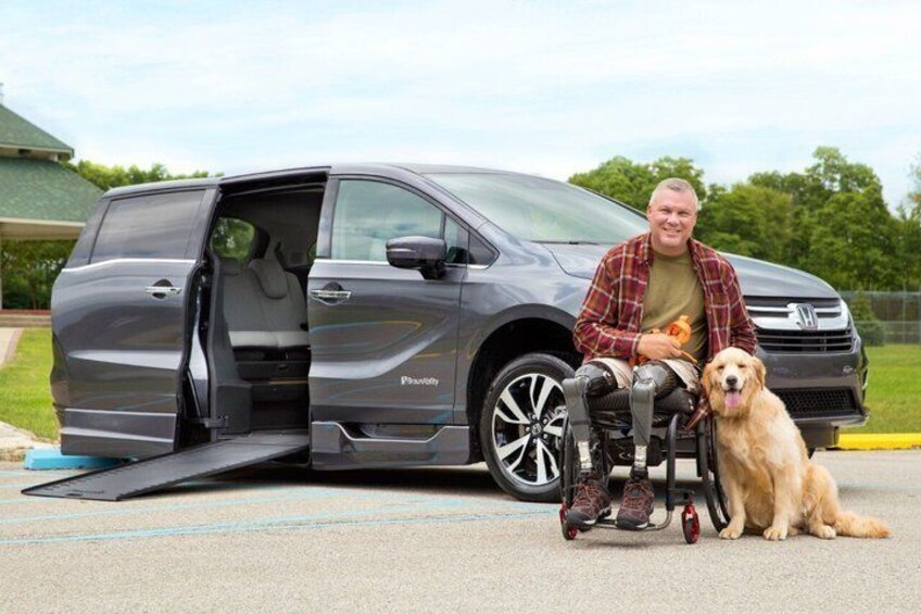 En person i rullstol står bredvid en skåpbil med en ramp, och en hund sitter bredvid personen. 1