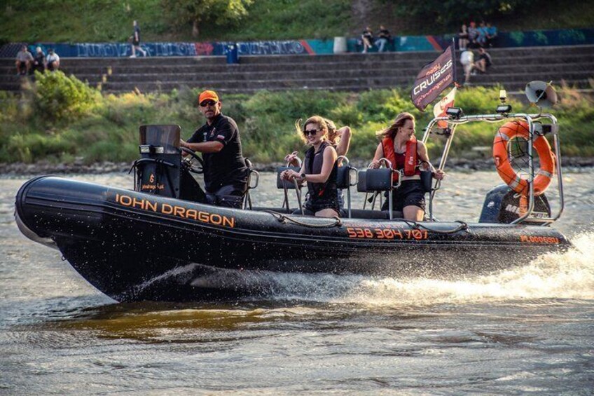 A black inflatable boat named 'John Dragon' with people on board, water spray, and a riverbank with spectators. 3