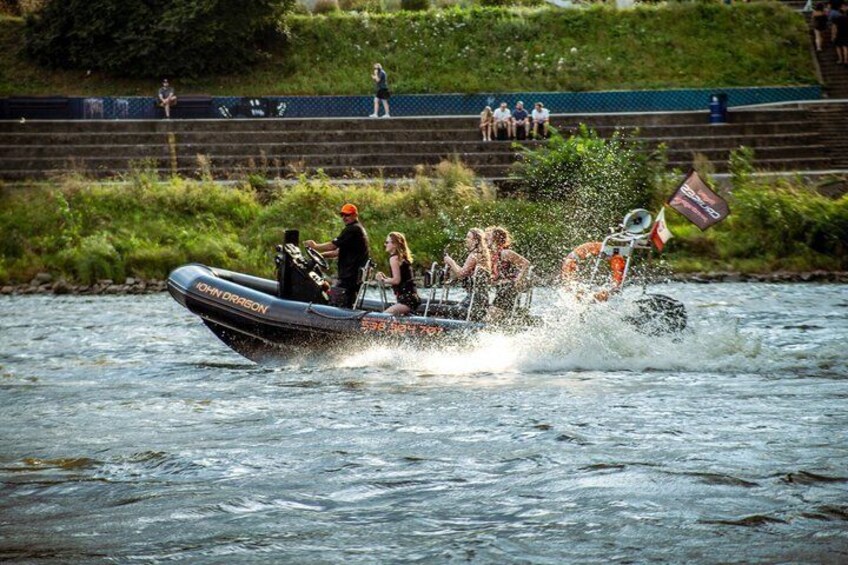 A speedboat with passengers, splashing water, on a river with spectators on the bank. 2