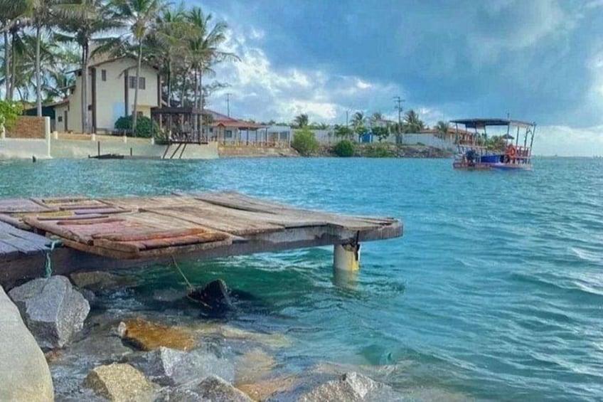 A wooden dock extending into calm blue water, a small boat in the distance, and a coastal building with a red roof. 1