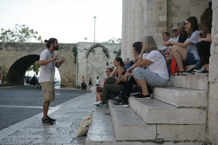 Un homme fait des gestes en direction d’un groupe de personnes assises sur des marches. 1