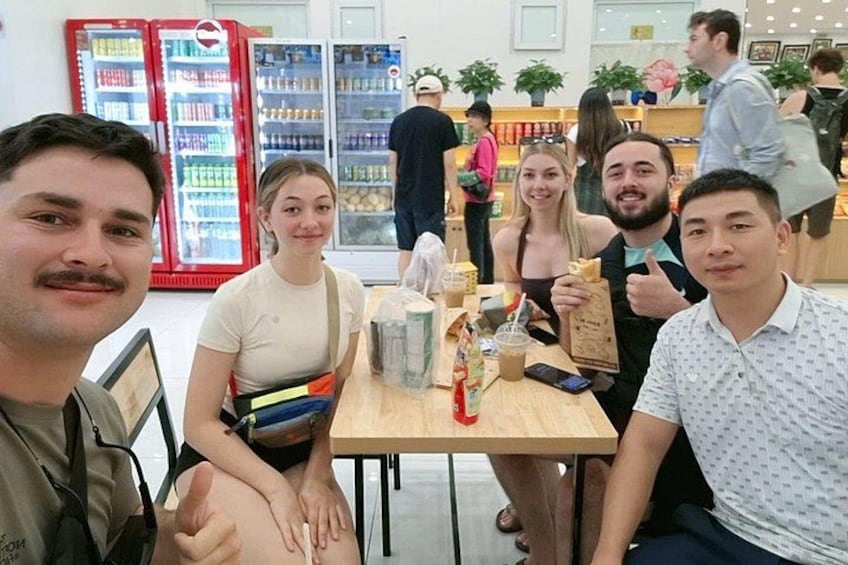 A group of people sitting at a table with drinks and a menu in a cafe with refrigerators in the background. 1