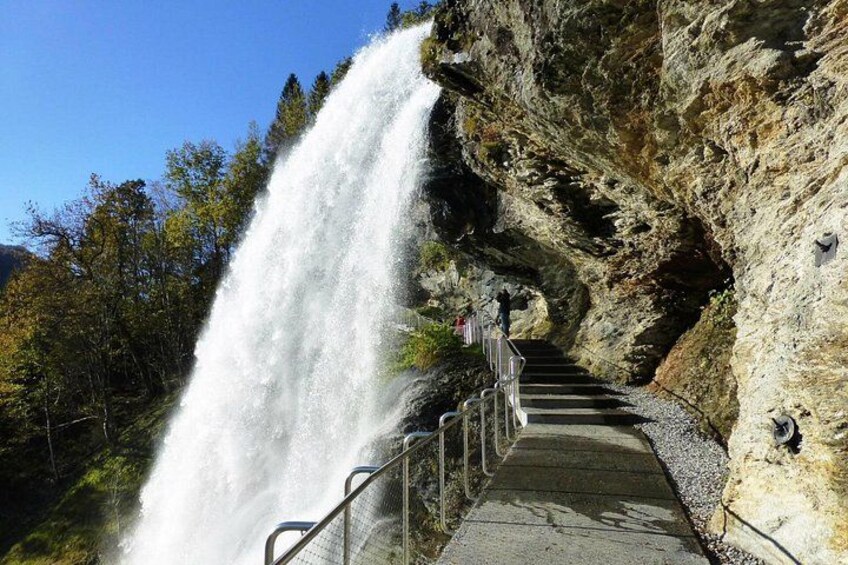 A waterfall cascading down a rocky cliff, a concrete pathway with metal railings, and a forested area in the background. 1