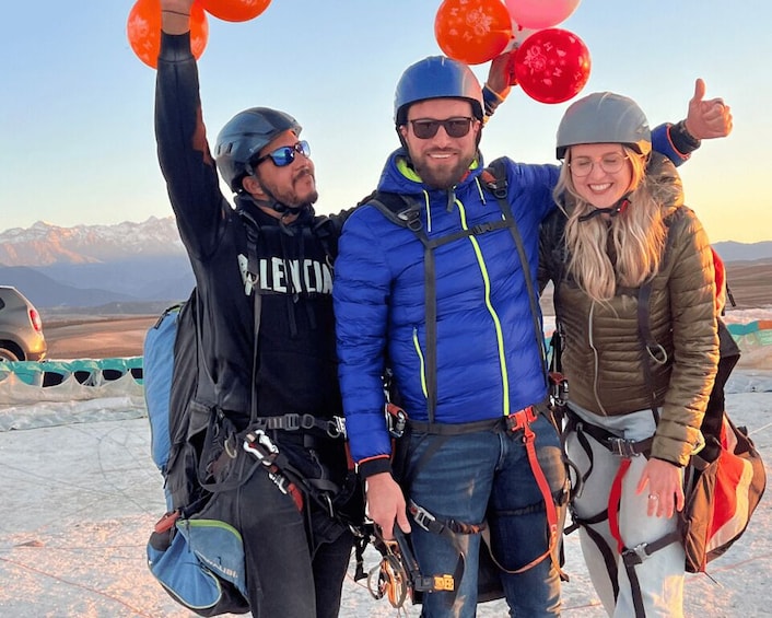 Three people in winter gear with helmets and harnesses, holding balloons, and standing on a snowy mountain with a clear sky. 5