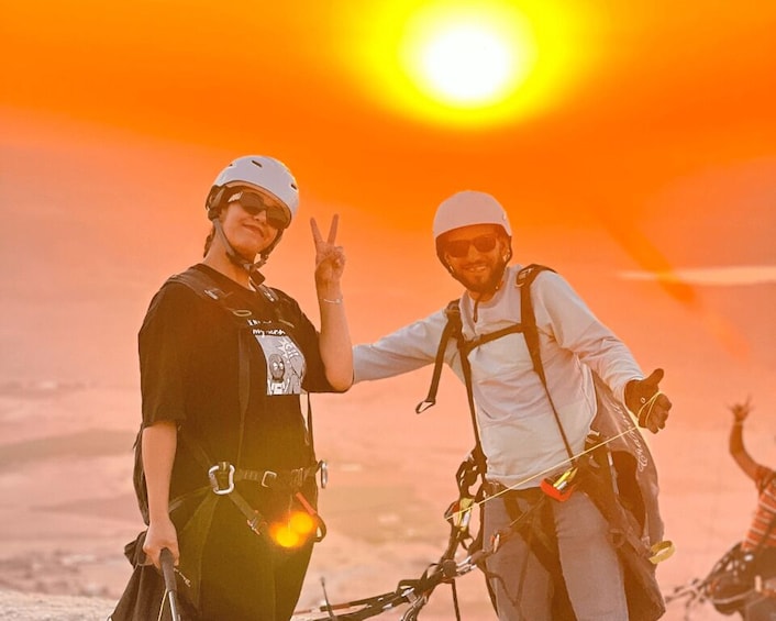 Two people with helmets and harnesses, one making a peace sign, against a sunset background. 3