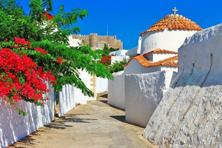 A narrow street with white-walled buildings, a church with a red-tiled roof, and a castle in the background. 2