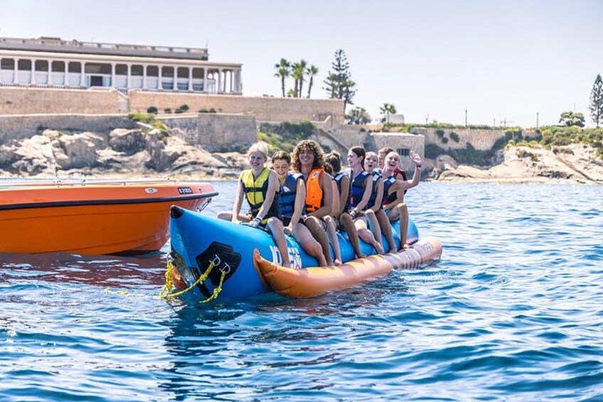 Un gruppo di persone su un barchino gonfiabile in acqua, con una barca e una costa rocciosa sullo sfondo. 1
