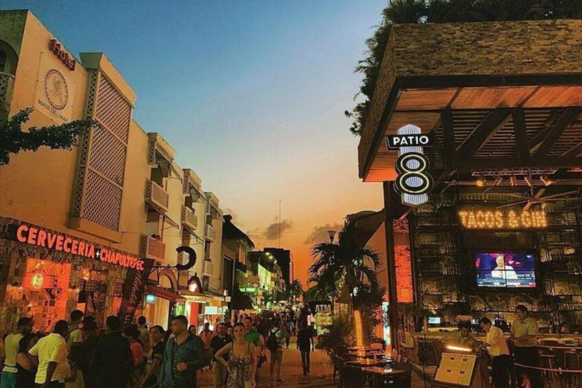 Una calle concurrida al anochecer, con tiendas, un bar y gente caminando. 4