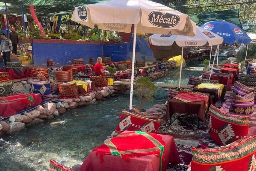 A riverside market with colorful boats and tables, and umbrellas with brand names. 1