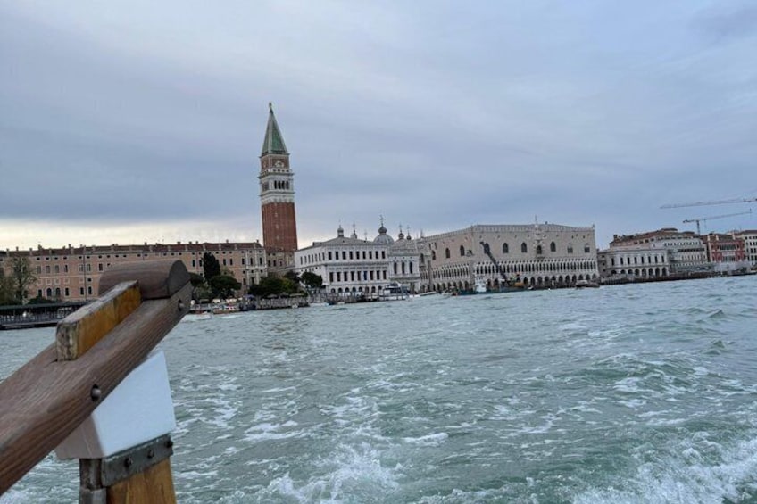 A river with a wooden structure in the foreground, a large building with a tower in the background, and a cloudy sky. 2