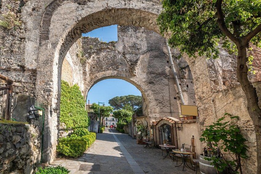 An ancient stone archway with a cobblestone path, greenery, and outdoor seating. 2
