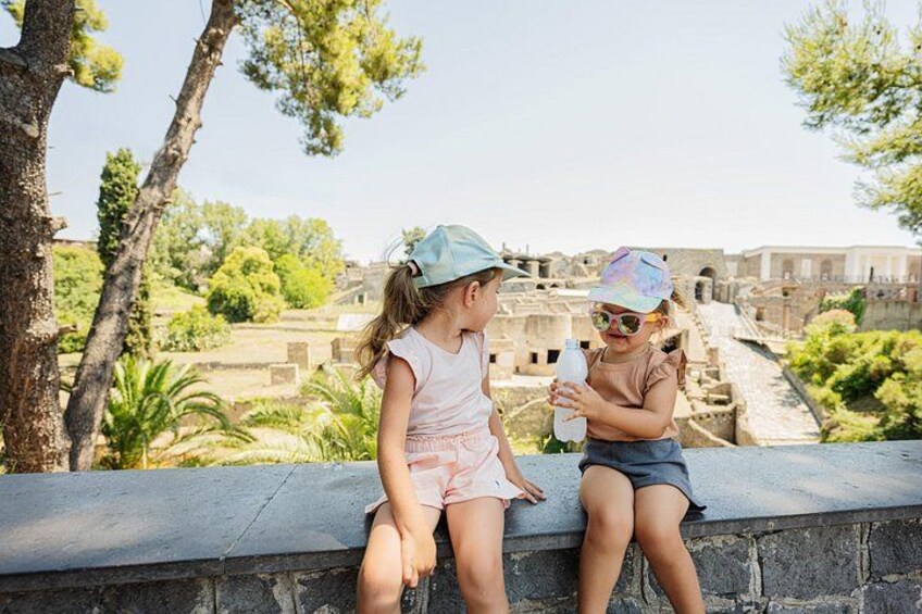 Two children sitting on a stone wall overlooking ancient ruins with trees and greenery in the background. 3