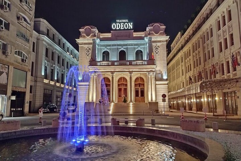 Ein prächtiges Gebäude, dessen Name 'ODEON' oben leuchtend zu sehen ist, umgeben von einem Springbrunnen und klassischer Architektur. 3