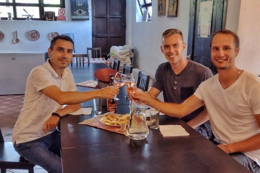 Drei Personen stoßen mit Weingläsern bei einem Tisch mit Essen und einer Flasche an. 2