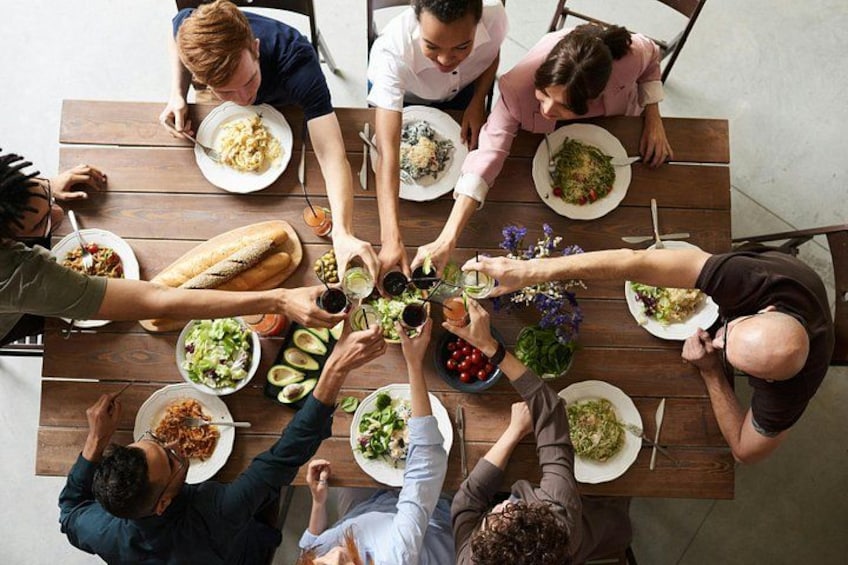 Eine Gruppe von Personen um einen Holztisch mit verschiedenen Speisen, darunter Salate, Brot und eine Weinflasche. 1