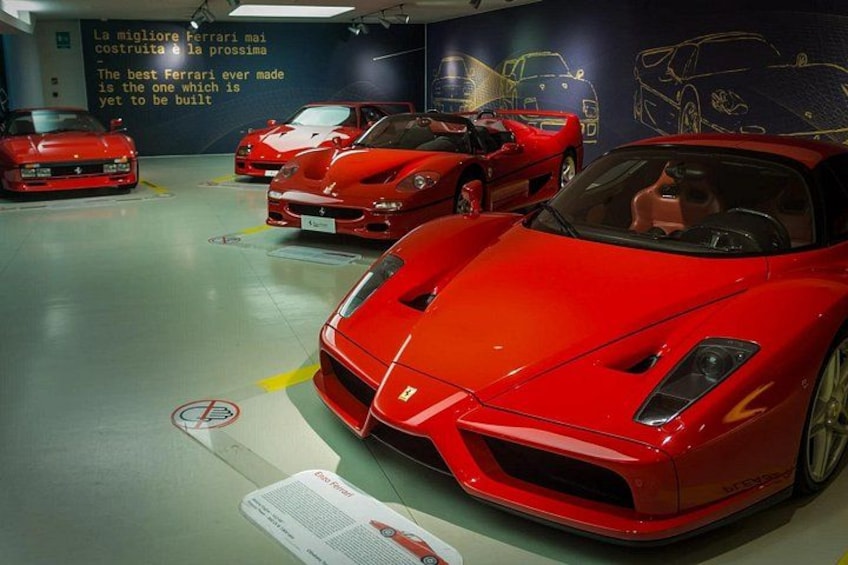A red Ferrari sports car in a showroom with other red cars and a wall with car drawings. 2