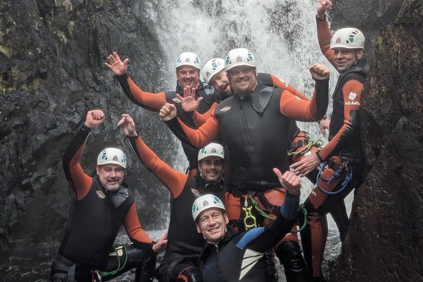 A group of people in black and orange wetsuits with helmets, standing in front of a waterfall. 2