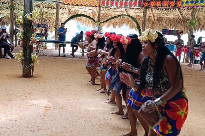 A group of people in colorful costumes, standing in a line under a thatched roof structure. 2