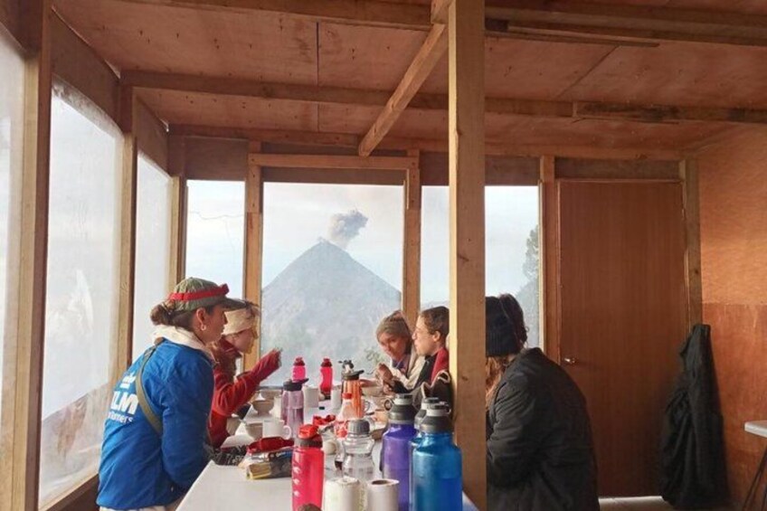 Un gruppo di persone sedute a un tavolo all'interno di una struttura in legno, con una montagna sullo sfondo. 2