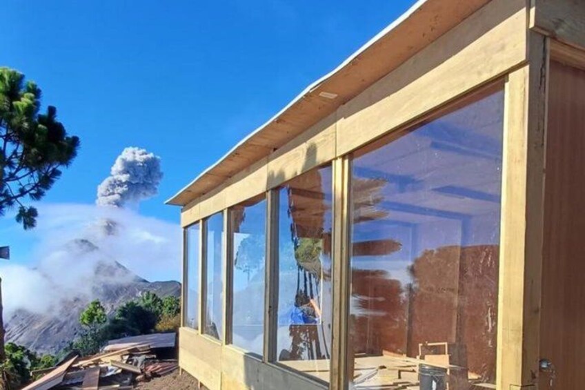 Un edificio moderno in legno con ampie vetrate, materiali da costruzione e una montagna sullo sfondo con una colonna di fumo. 3